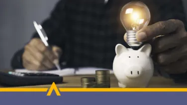 kobieta siedzącą przy stole z kalkulatorem i dokumentami, obok znajdują się skarbonka w kształcie świnki i żarówka